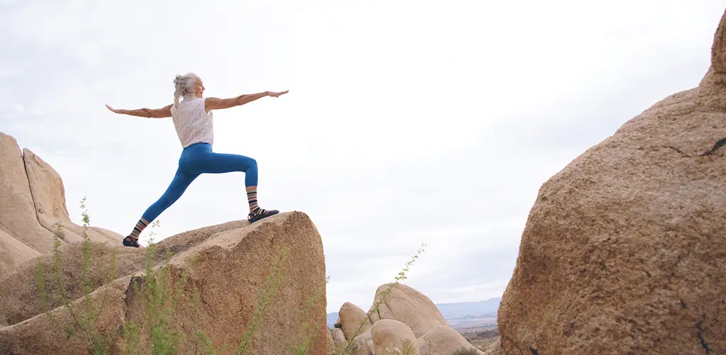 Eine ältere Frau in blauen Leggings praktiziert Yoga in einer felsigen Küstenlandschaft mit freiem Blick aufs Meer. Sie hält eine ausbalancierte Pose mit ruhiger Konzentration, umgeben von Klippen und offenem Himmel.