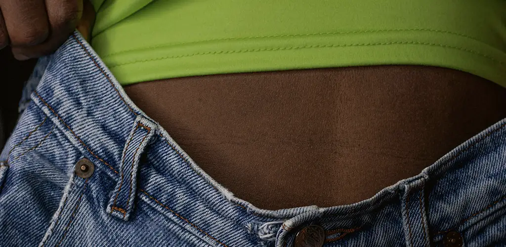 Person iført blå denimjeans trækker linningen væk fra maven, med en grøn top synlig.