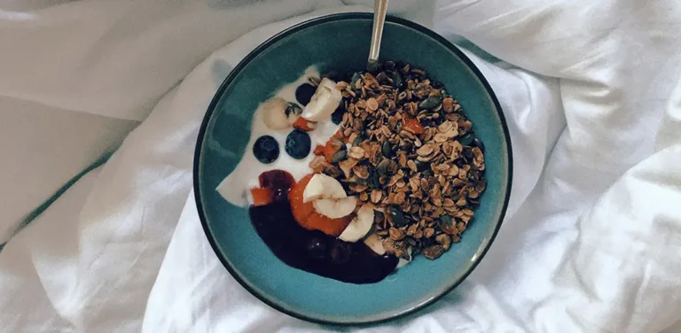 A bowl of yogurt topped with granola, banana slices, blueberries, and fruit jam, served with a spoon on a white bed sheet.