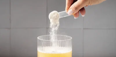 En skefuld proteinpulver hældes med hånden i et glas gul drik, mens en anden skefuld pulver placeres ved siden af ​​glasset på en hvid overflade.
