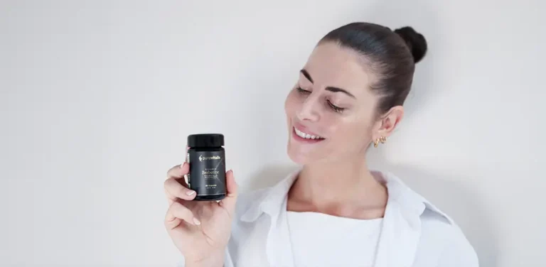 Eine lächelnde Frau mit dunklem, zu einem Dutt gebundenem Haar, die ein weißes Hemd trägt, hält ein black Eine Flasche Purovitalis Berberine Balance vor hellem Hintergrund.