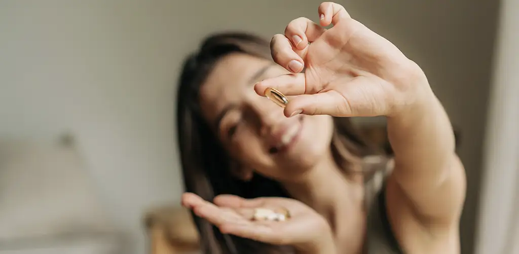 A smiling woman holding dietary supplements in her hand, showing a capsule between her fingers, symbolizing daily health and wellness.