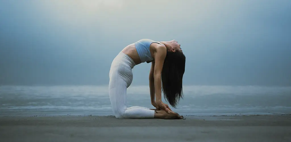 Una mujer practica yoga en la postura del camello (Ustrasana) en una playa al amanecer, con un top azul claro y unos leggings blancos