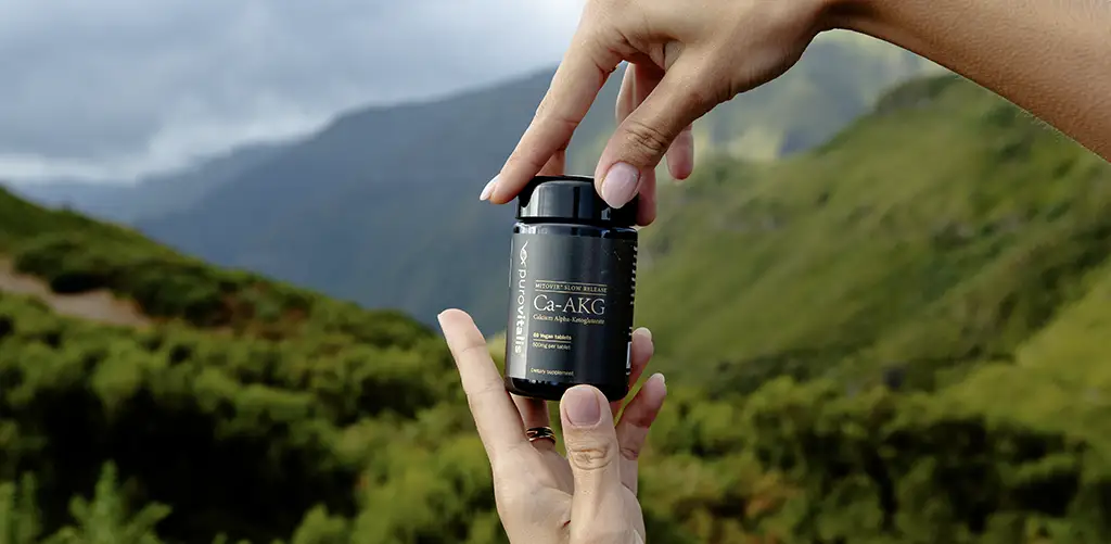 Mains tenant une bouteille de supplément Purovitalis Ca-AKG à l'extérieur avec des montagnes vertes en arrière-plan.