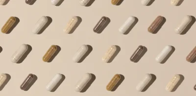 Supplements capsules arranged in a neat repeating pattern on a beige background, in varied shades of white, tan, and brown, casting soft shadows.