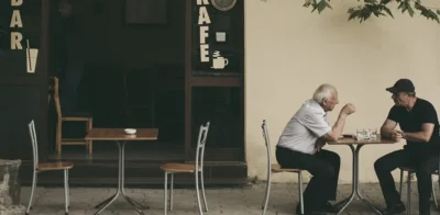 To ældre mænd sidder ved et lille udendørs cafébord og er engagerede i en samtale, hvilket illustrerer social forbindelse og kammeratskab senere i livet.