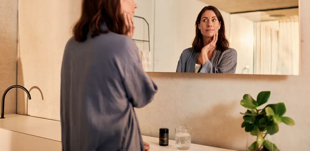 Eine Frau steht in einem Badezimmer, berührt sanft ihr Gesicht und betrachtet ihr Spiegelbild; auf dem Waschbecken steht das Produkt Purovitalis Resveratol.